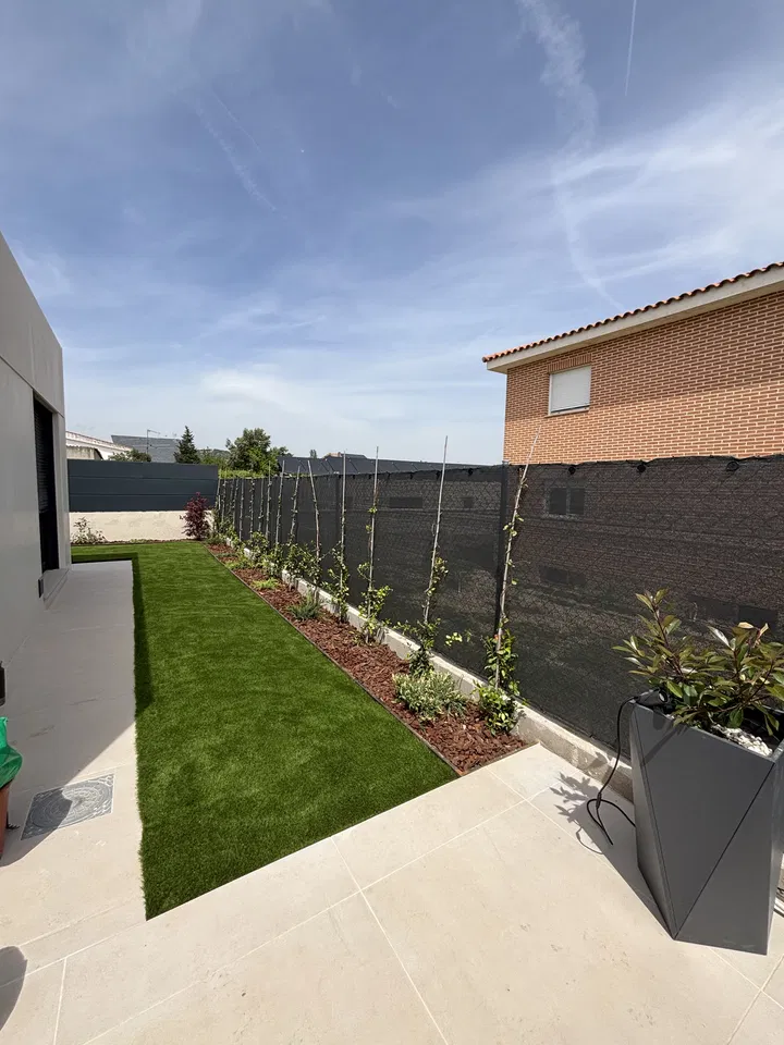 Jardín residencial alargado con césped, plantaciones junto a una valla y pavimento; fotografía 1 de esta serie