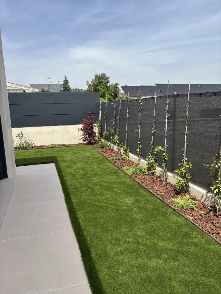 Jardín residencial alargado con césped, plantaciones junto a una valla y pavimento; fotografía 3 de esta serie