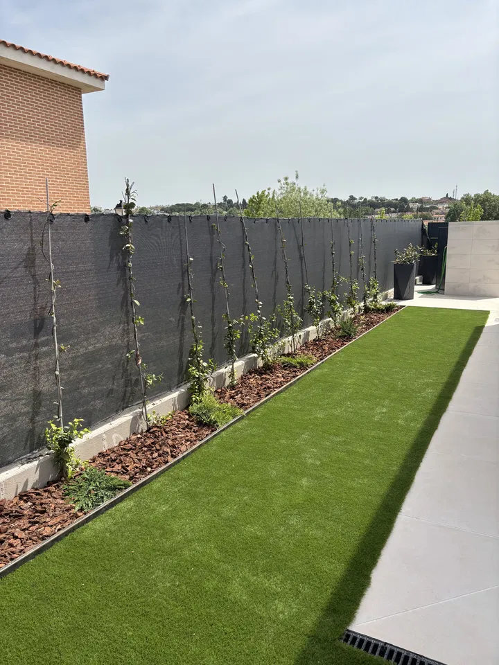 Jardín residencial alargado con césped, plantaciones junto a una valla y pavimento; fotografía 5 de esta serie