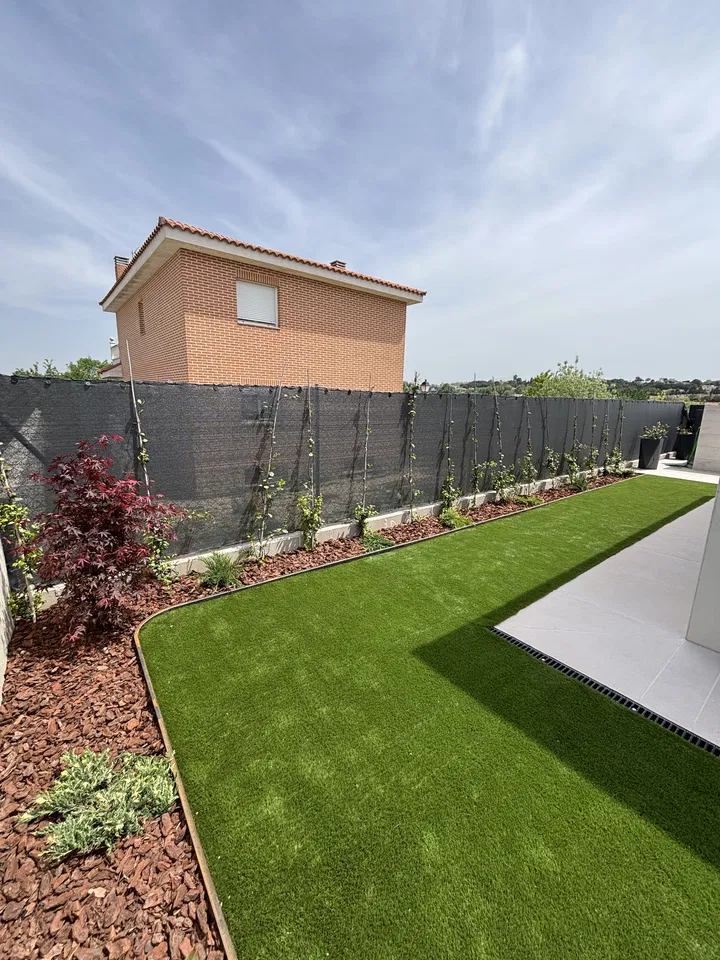 Jardín residencial alargado con césped, plantaciones junto a una valla y pavimento; fotografía 7 de esta serie